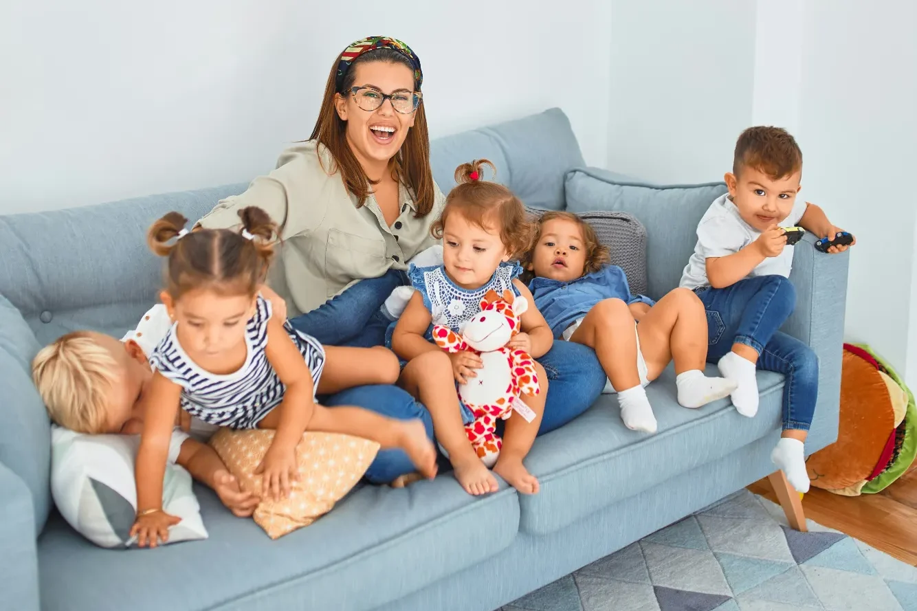 Eine fröhliche Frau sitzt mit fünf Kindern auf einem grauen Sofa.