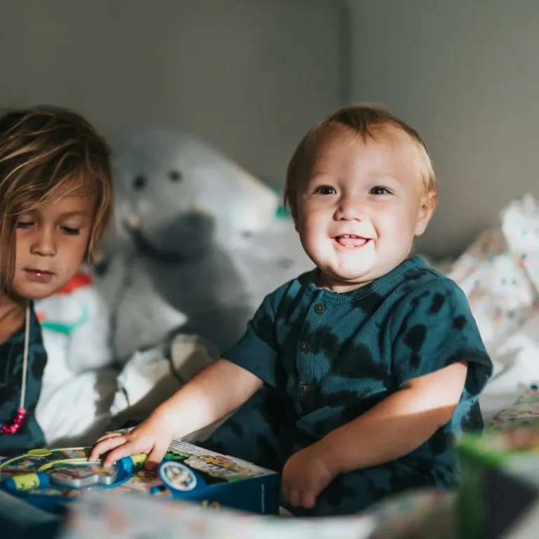 Zwei Kinder spielen im Zimmer, eines lächelt und hält Spielzeug in der Hand.