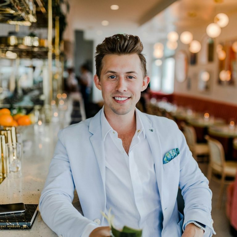 Lächelnder Mann in einem hellen Sakko, sitzt an einer Bar mit Getränk in der Hand.