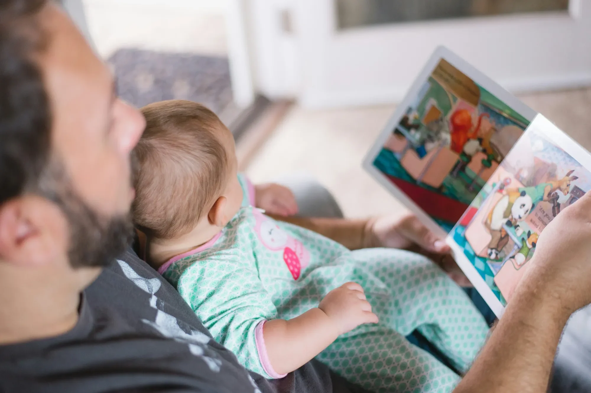 Vater liest einem Baby ein Bilderbuch vor, beide entspannt im Wohnraum.
